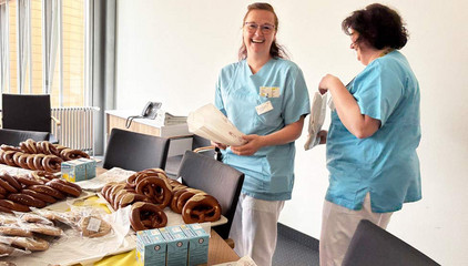 Zwei Pflegekräfte stehen in blauer Funktionskleidung um ein Buffet zum Internationalen Tag der Pflegenden in der Immanuel Klinik Rüdersdorf und unterhalten sich fröhlich.