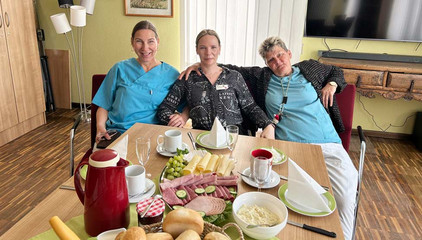Drei Pflegekräfte in Funktionskleidung sitzen nebeneinander auf einem Sofa an einem gedeckten Frühstückstisch und blicken in die Kamera beim Internationalen Tag der Pflegenden im Immanuel Haus am Kalksee in Rüdersdorf bei Berlin.